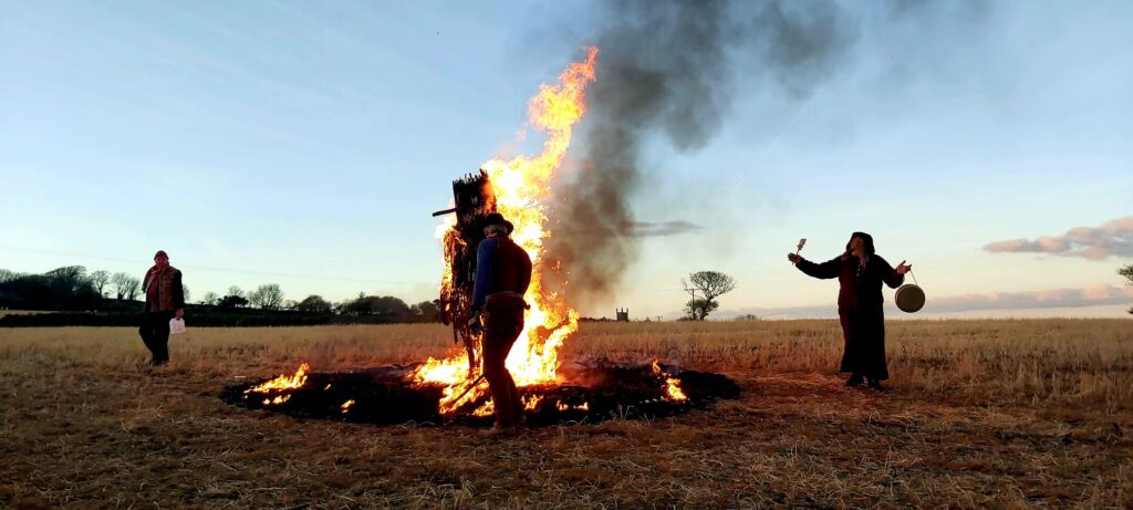 a ritual figure is burned in an open field, against a blue sky while human figures move around it