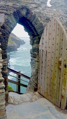 antique stone doorway with pointed peak and wooden door opened to see a rugged coastline with deep blue sea and surf